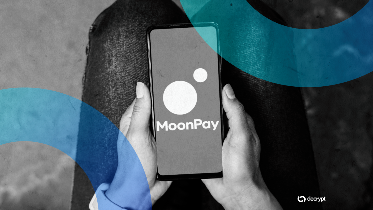 Person holds a smartphone displaying the MoonPay logo in a grayscale photo with blue circular overlays and the Decrypt logo bottom right.