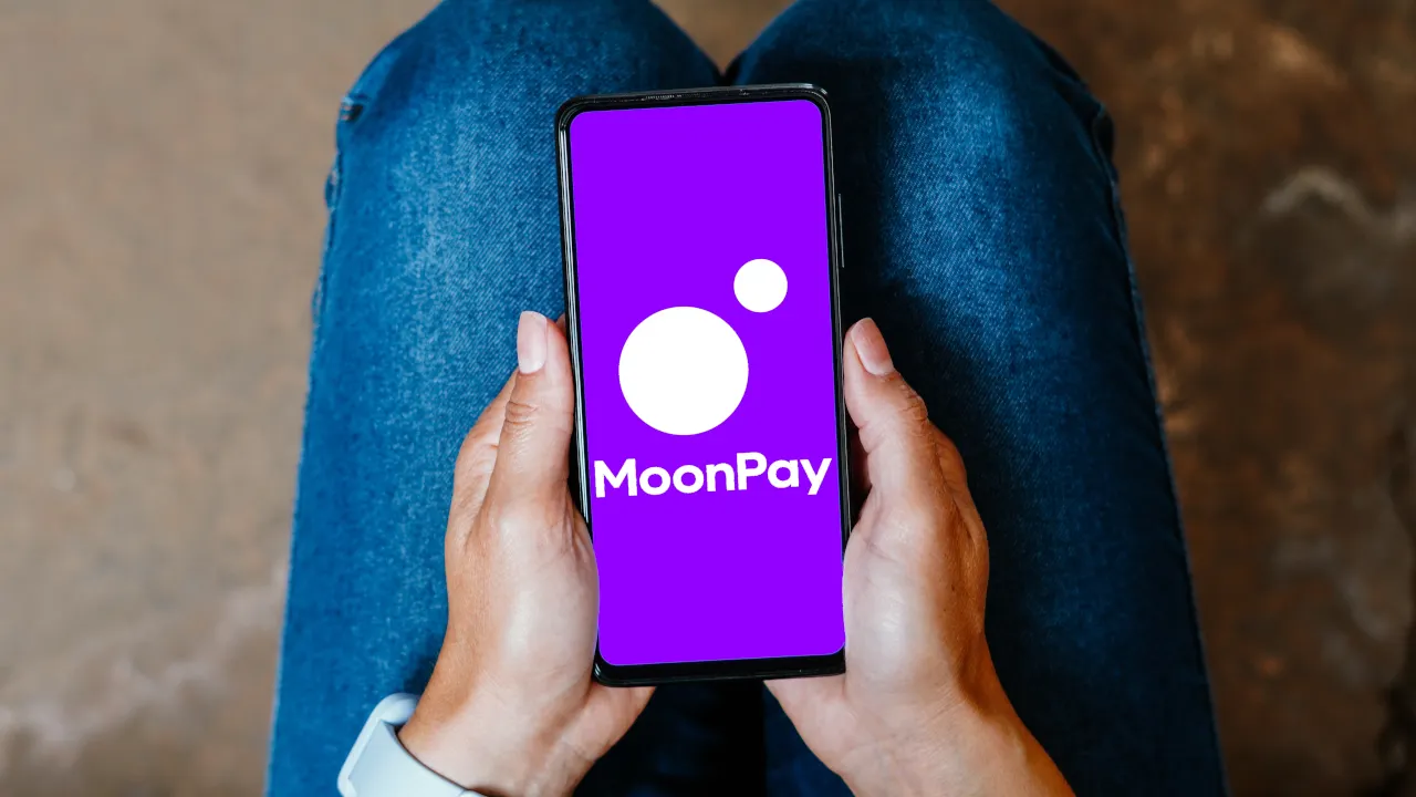 MoonPay app loaded on a smartphone screen held by both hands, with a purple background and white logo.