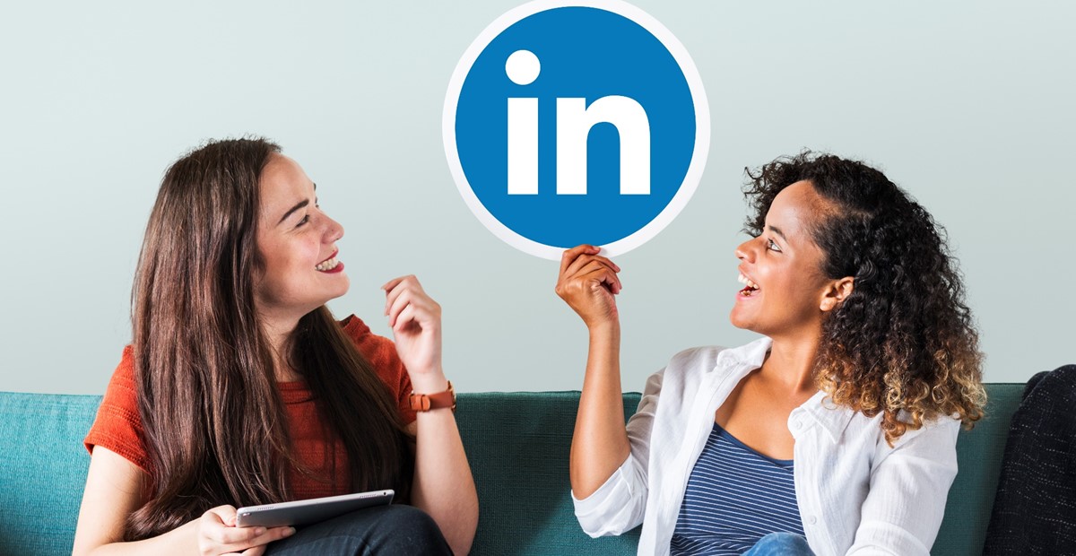 Two women sit on a sofa, smiling at each other, one holding a tablet while a large LinkedIn logo is between them.